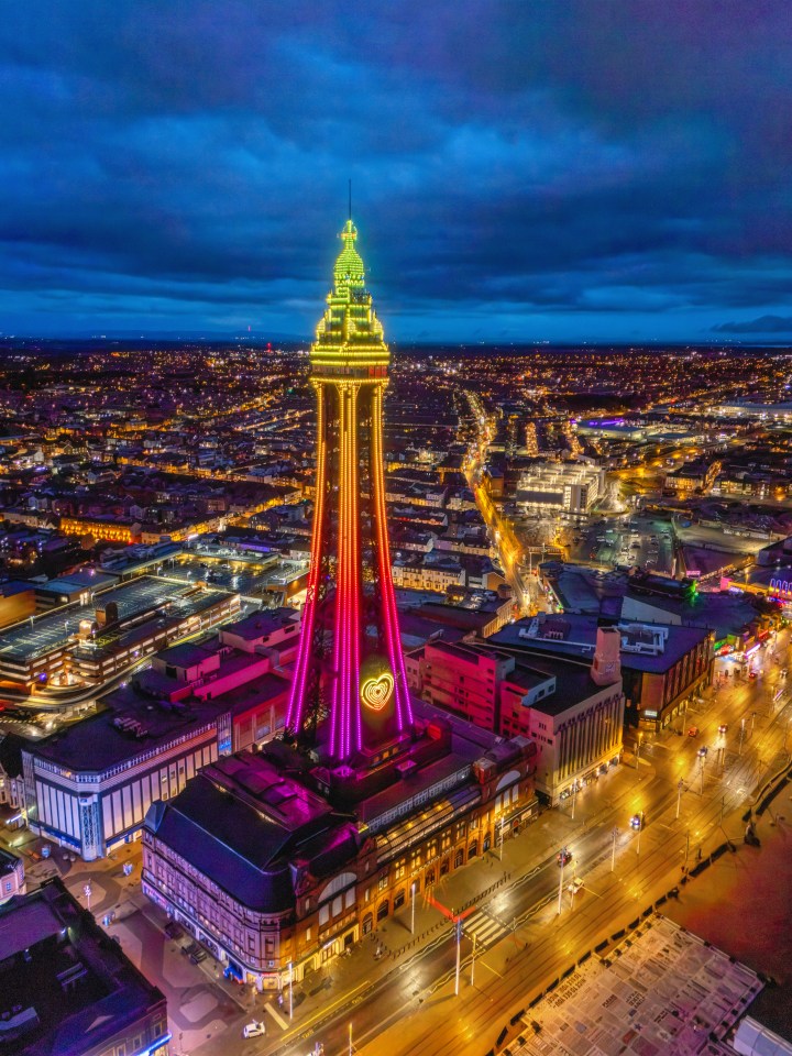Aerial view of Blackpool Tower at night, illuminated in yellow, red, and pink lights, with a yellow heart symbol on its lower section, surrounded by the illuminated city.
