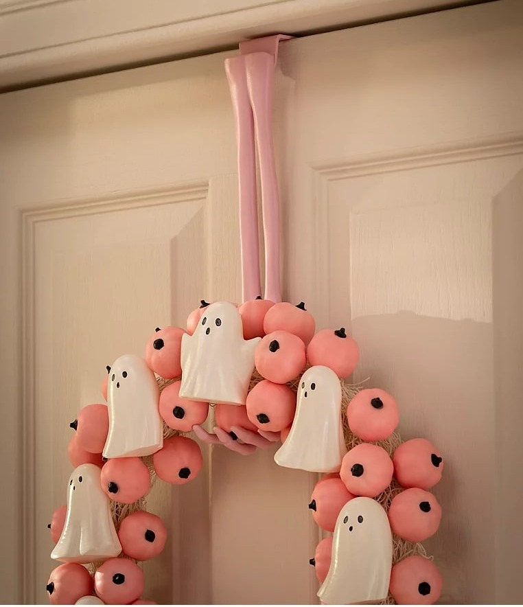 Halloween wreath made of small pink pumpkins and white ghosts, hanging on a white door.