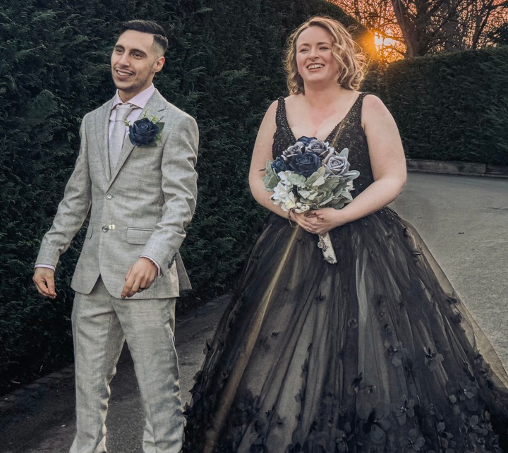 A groom in a light grey suit with a dark blue boutonnière, standing next to a bride in a black wedding dress, holding a bouquet of dark flowers, with a sunset sky in the background.