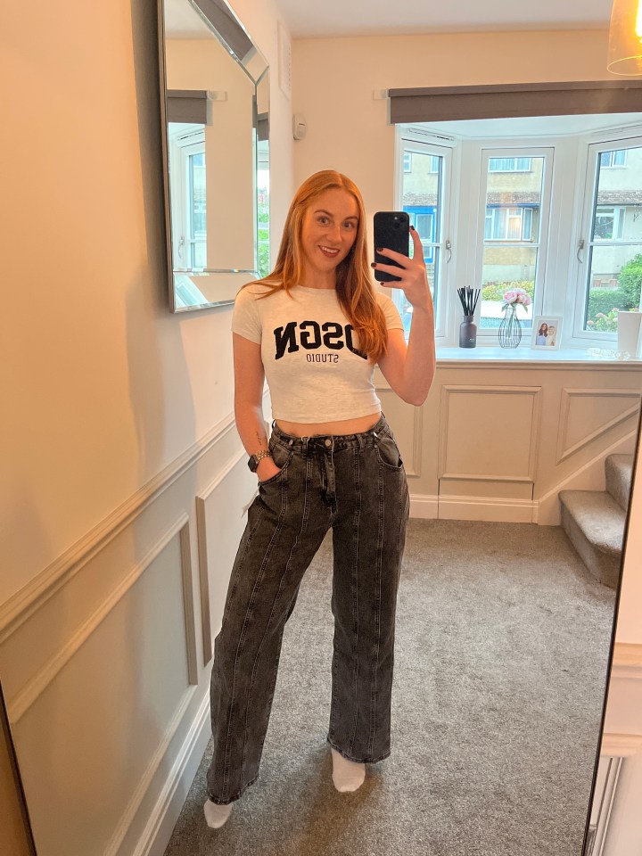 A woman in a white t-shirt and grey jeans takes a mirror selfie.