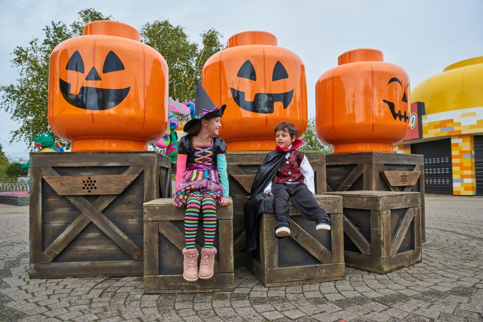 Two children in Halloween costumes sit on wooden crates in front of large Lego brick jack-o'-lanterns at Legoland.