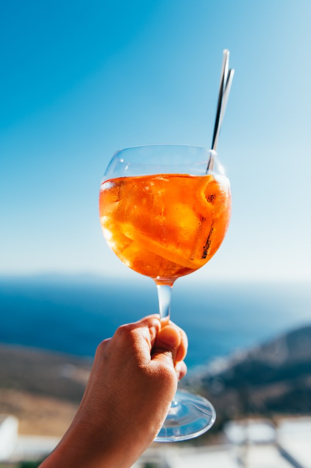 Spritz cocktail with ice and orange slices in a wine glass held against a blue sky and sea background.