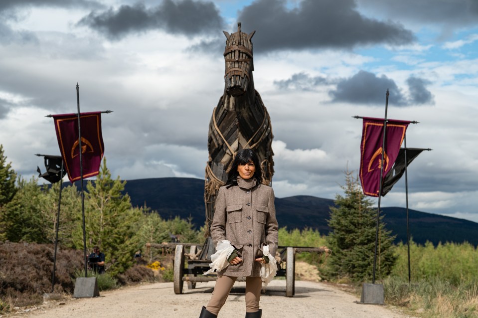 A presenter in a brown coat stands in front of a large wooden Trojan horse-like structure, flanked by two flags, with mountains and pine trees in the background.