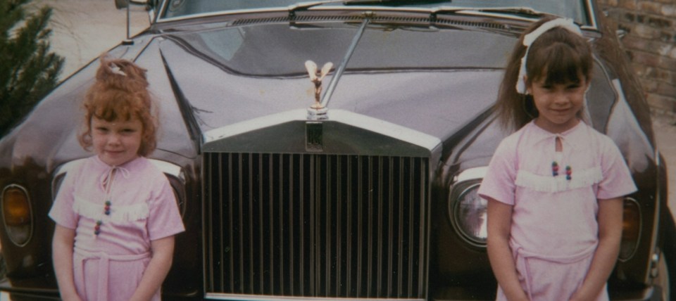 Two young girls in pink dresses standing in front of a dark luxury car.