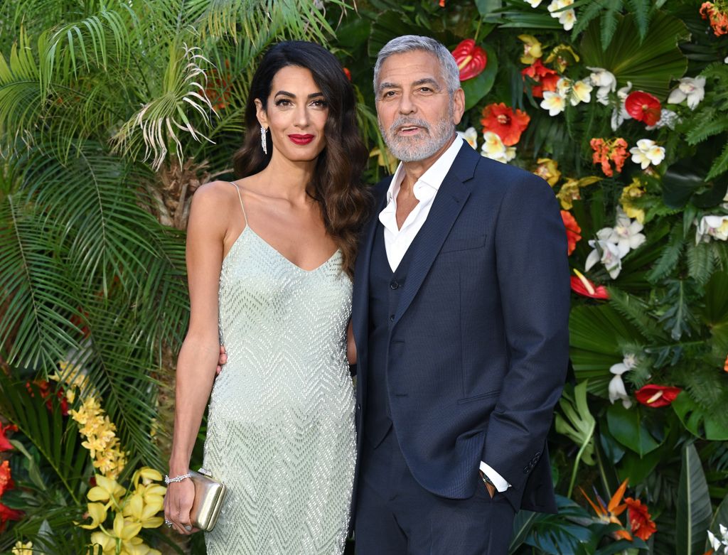 George Clooney and Amal Clooney stand together in front of plants and tree backdrop while attending film premiere