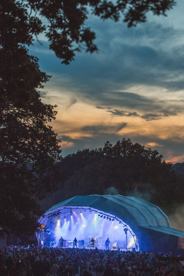 Outdoor concert stage with a band performing under blue lights to a large crowd at dusk.