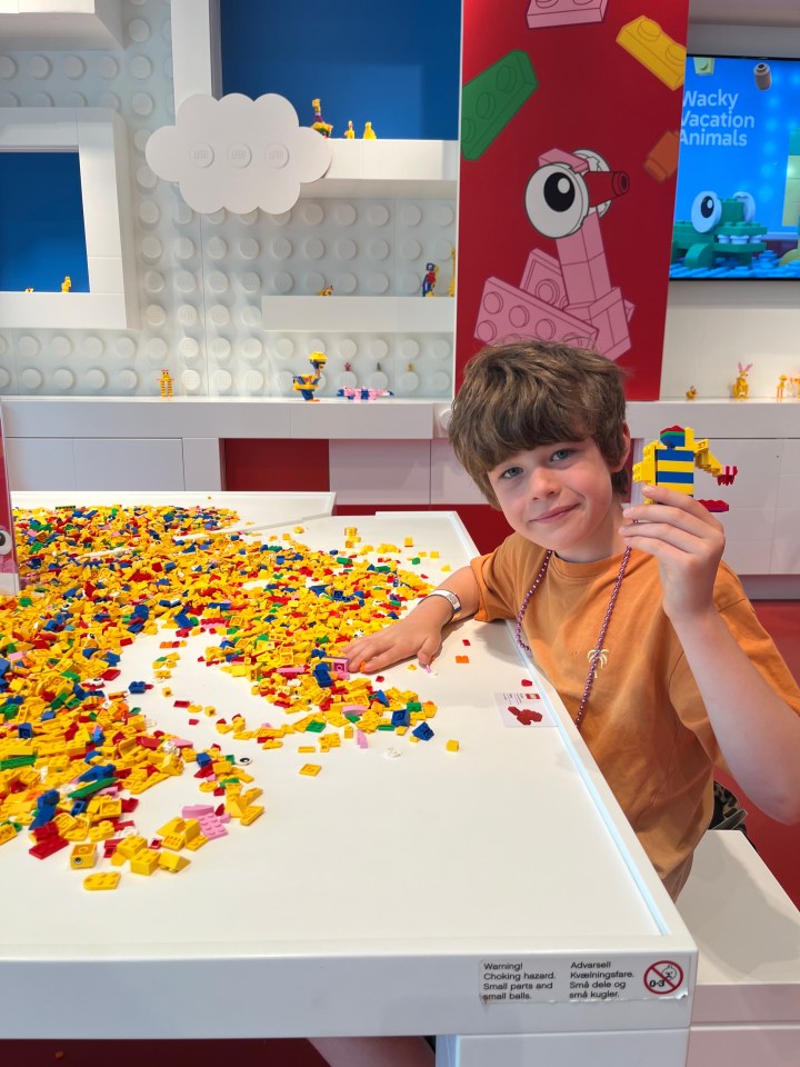 A boy holding a yellow and blue Lego creation while sitting at a table covered in Lego bricks.