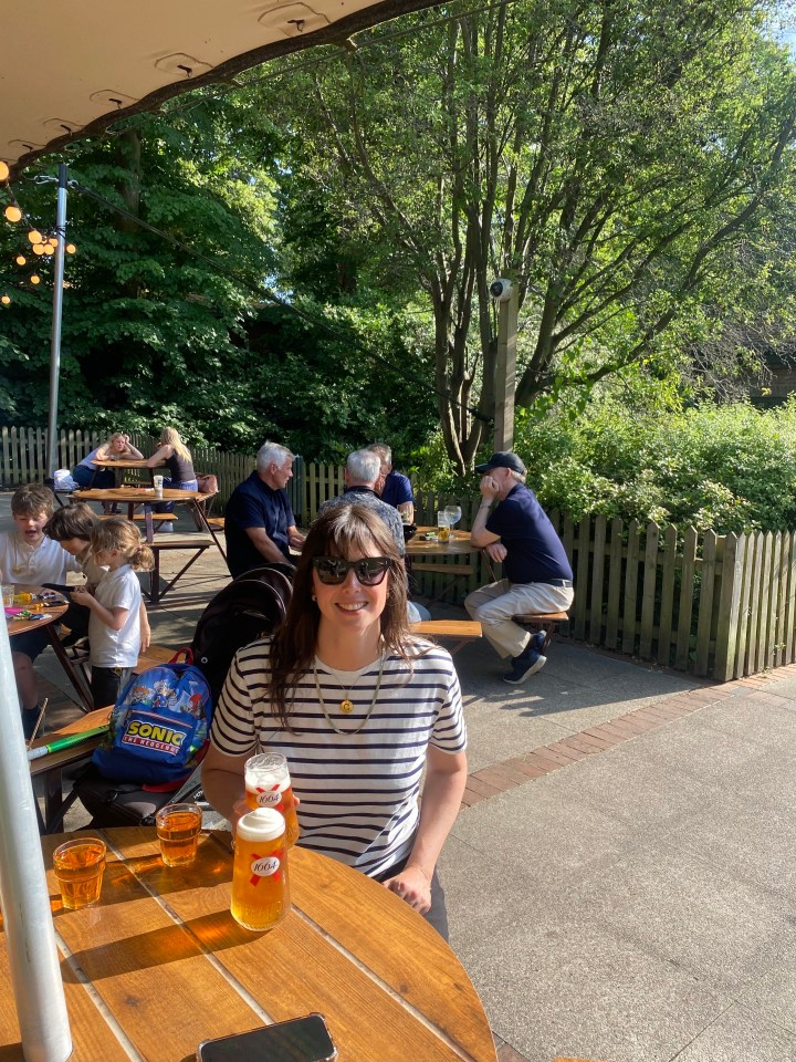 A woman holding two glasses of beer smiles at the camera while sitting at an outdoor restaurant.