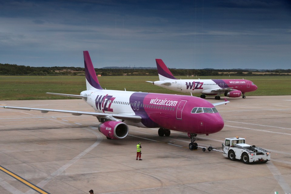 Two Wizz Air Airbus A320 aircraft, one being towed by a vehicle, on an airport tarmac with a person standing nearby.