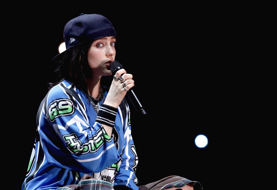 Billie Eilish performing onstage in a blue and white patterned shirt and a dark baseball cap, holding a microphone to her mouth.