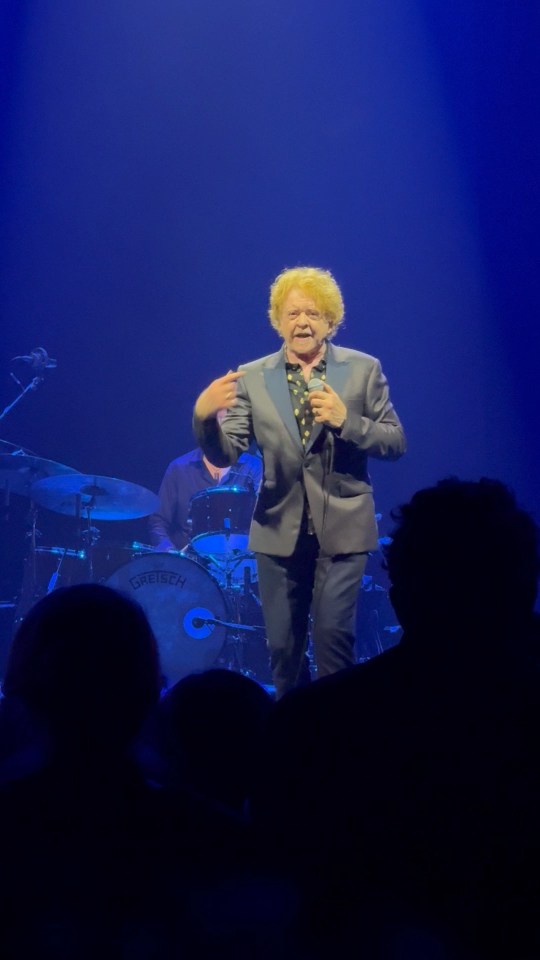 Mick Hucknall of Simply Red performing on stage, wearing a suit and holding a microphone, with a drummer visible behind him.