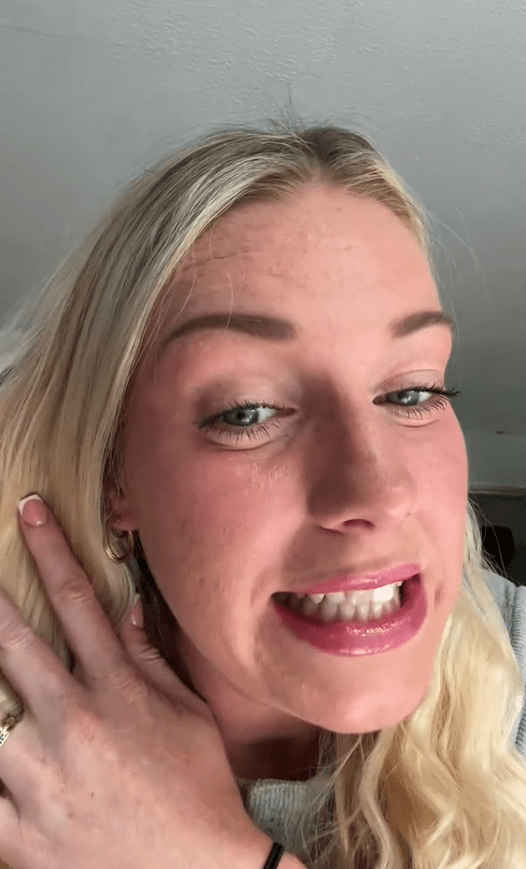 A young woman with blonde hair, red cheeks, and pink lipstick looks at the camera with her hand near her ear.