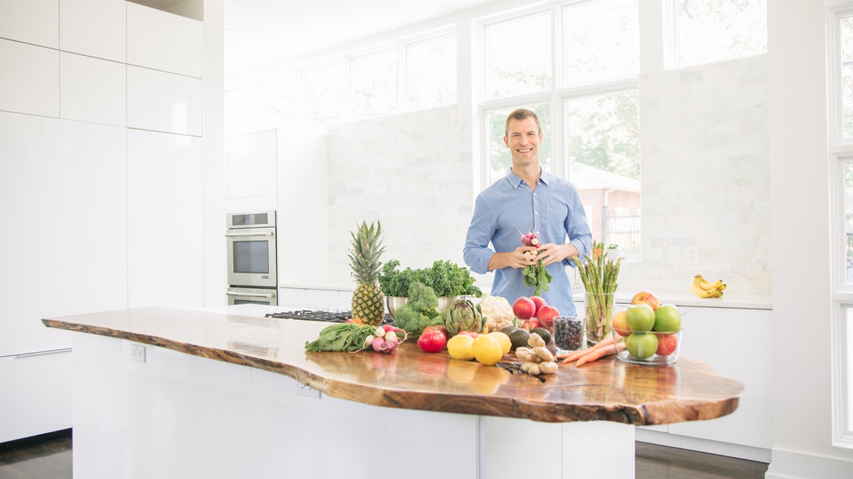 Dr. Axe standing in a white kitchen with a table covered in fruits and vegetables