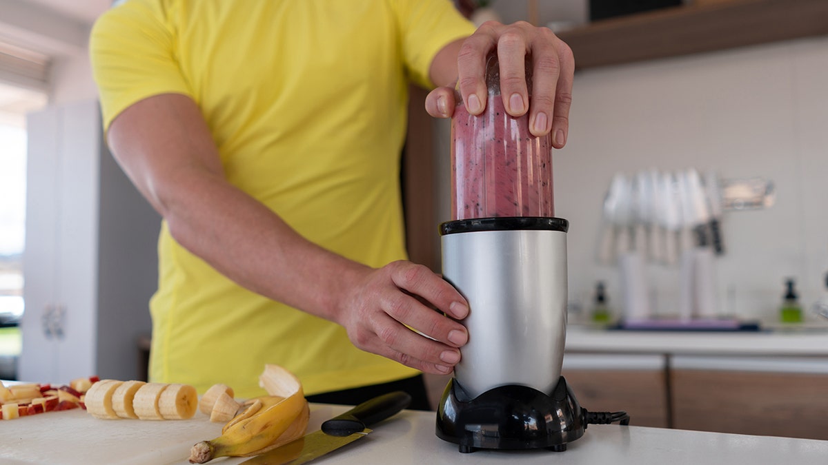 Close-up of a man in kitchen making a healthy smoothie for breakfast. Knife and banana seen on cutting board as he puts small blender together.
