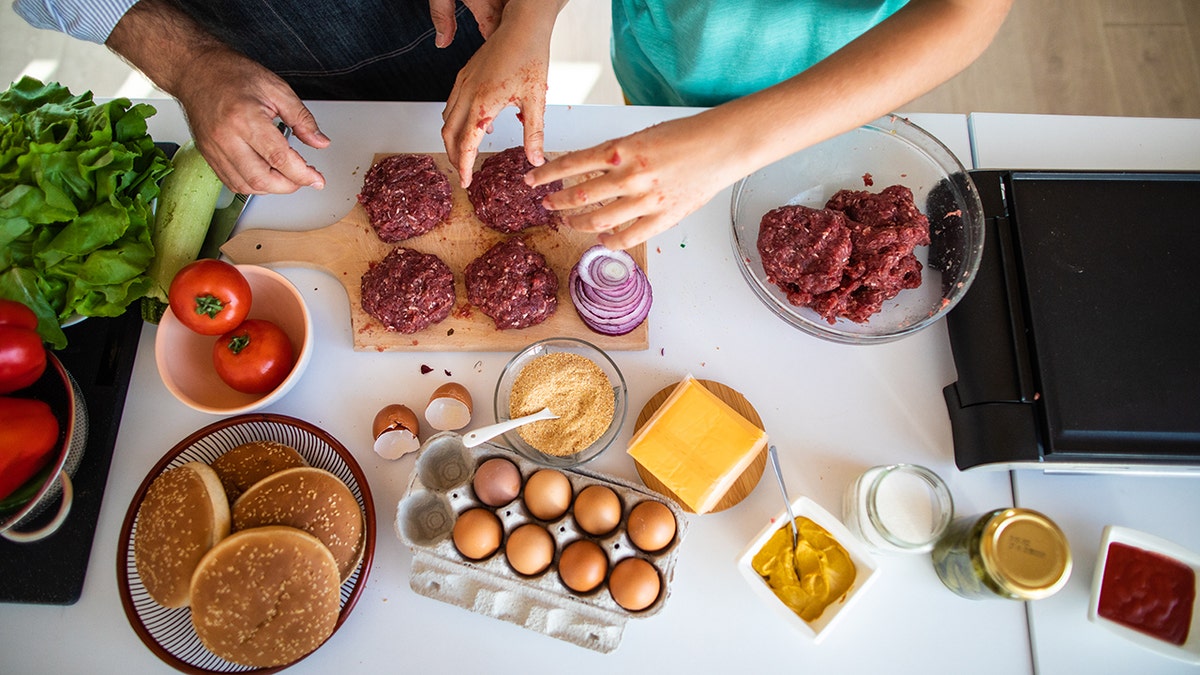 Father and son making cheeseburgers in kitchen, hands seen with ingredients including eggs, cheese, buns and chopped meat spread near griddle, indicating healthy cysteine-packed diet.