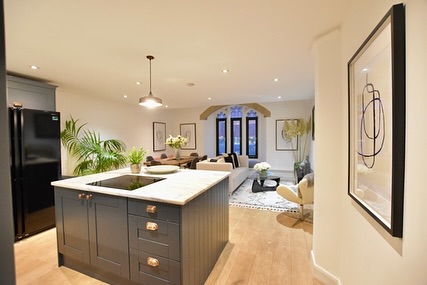 Katherine Ryan's kitchen and living area, featuring a dark kitchen island, black refrigerator, light wooden flooring, and a stone archway leading to the living room with large gothic-style windows.