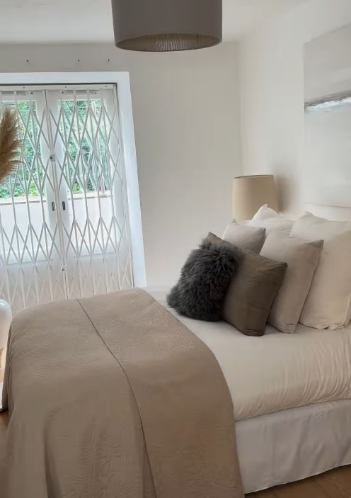 A white bedroom with a bed covered in a tan duvet and multiple pillows, and a window with a retractable security gate.