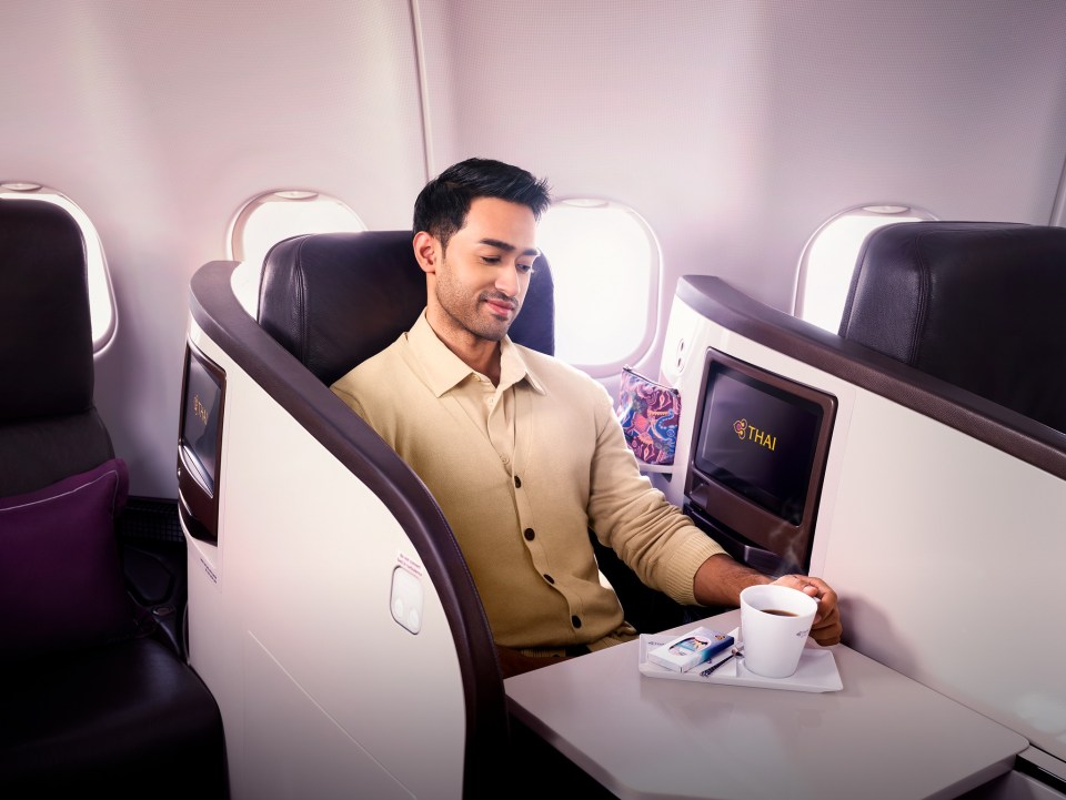 A man sits in a lie-flat premium economy airplane seat, holding a cup of coffee.