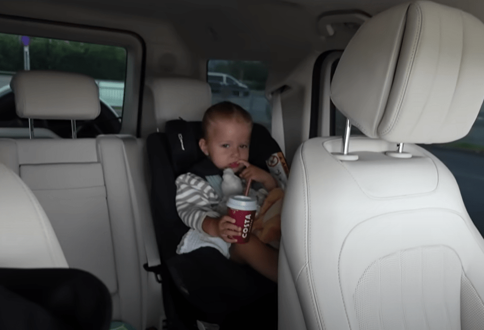 A child sits in a car seat in the back of a car, holding a red Costa cup with a straw.