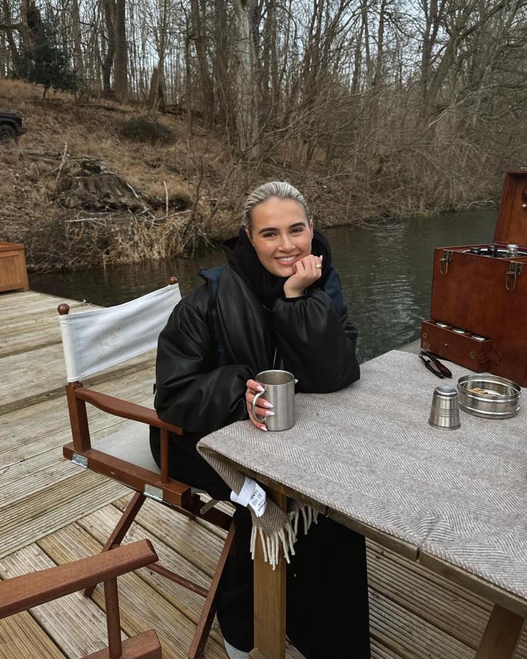 Molly-Mae sitting at a table with a metal cup next to a river with bare trees in the background.