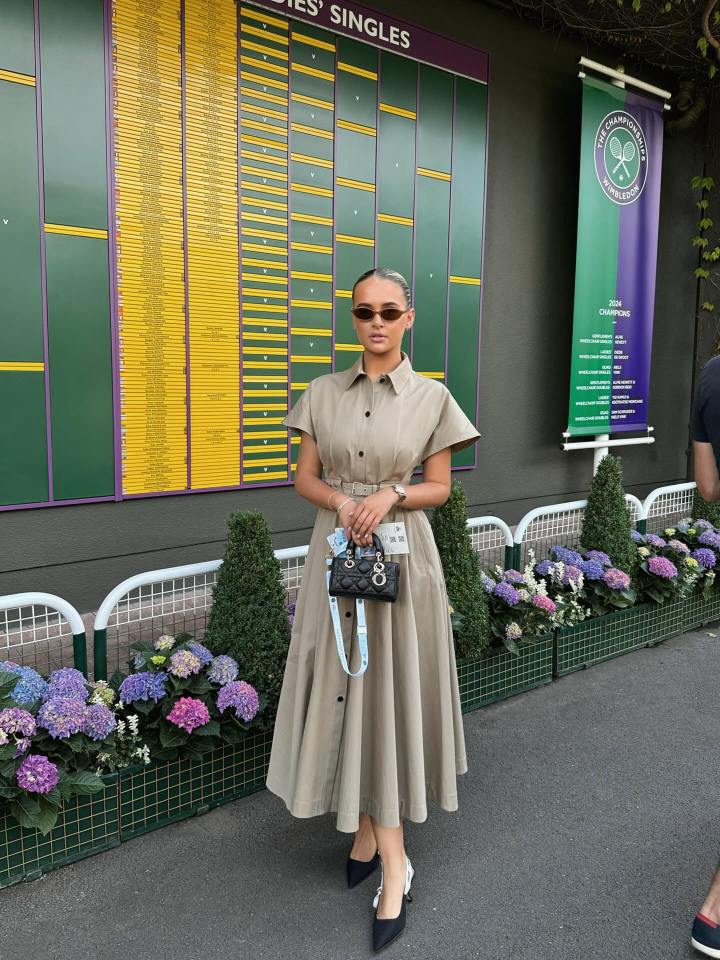 Molly Mae at Wimbledon in a belted tan dress, holding a small black purse, standing next to a score board.