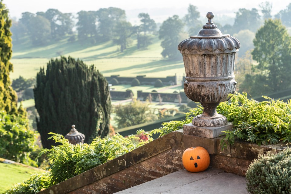 Jack-o'-lantern on a stone staircase with a large garden urn and landscaped gardens in the background.