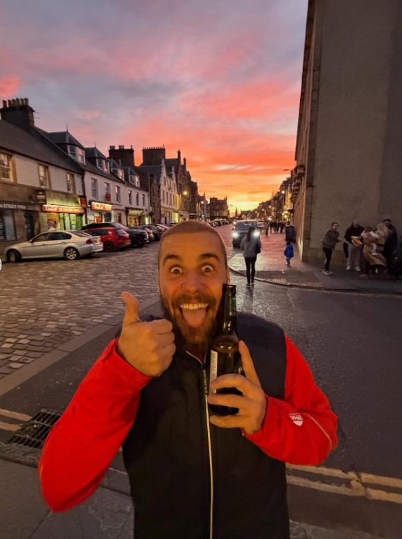 Justin Bieber holding a bottle of Innis & Gunn beer, sticking his tongue out, and giving a thumbs up at sunset in Scotland.