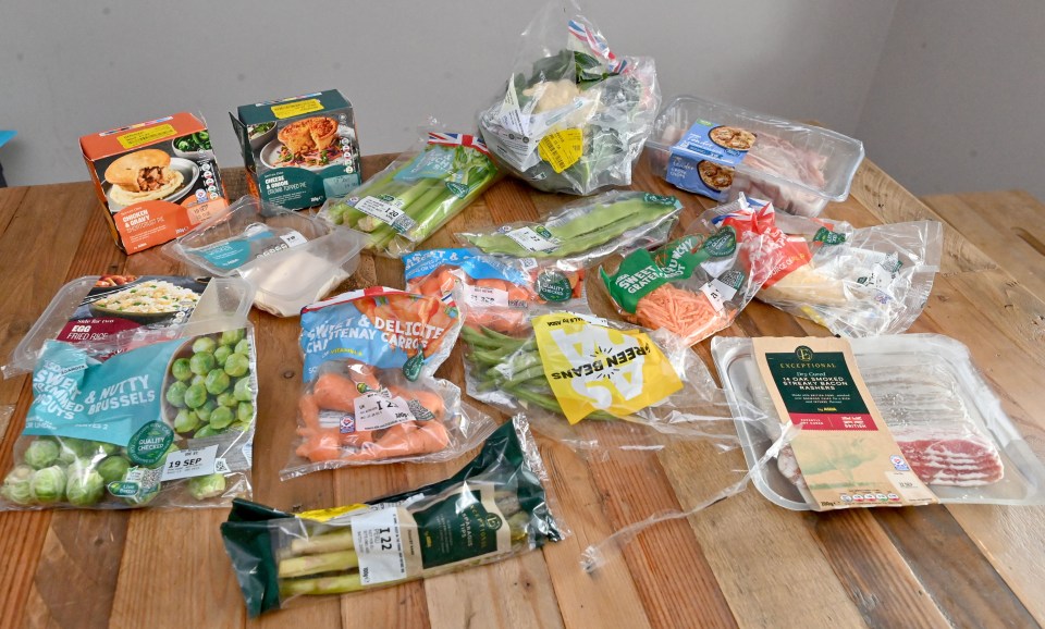 An assortment of packaged foods, including meat, vegetables, and pre-made meals, many with "reduced" stickers, are displayed on a wooden table.