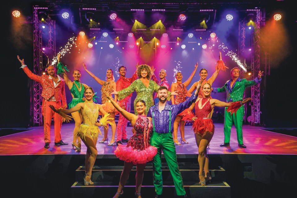 A group of performers in dazzling, colorful outfits on a stage with bright lights and pyrotechnics.