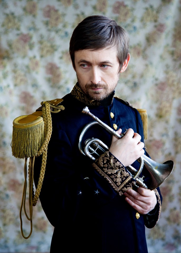 Man with a goatee in a navy military uniform with gold trim and epaulets, holding a silver trumpet.