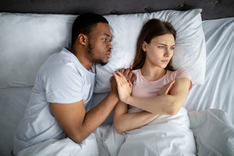 An African American man in bed trying to comfort a white woman who looks away with her arms crossed, refusing him.