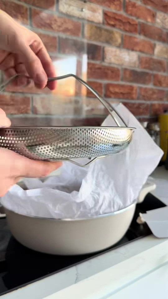 A person's hands holding a metal colander with a handle over a beige pot lined with white parchment paper.