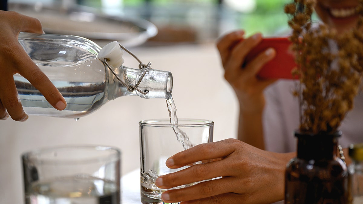 Waiter pouring water for customer at restaurant, who is smiling and filming or photographing the process on their phone.