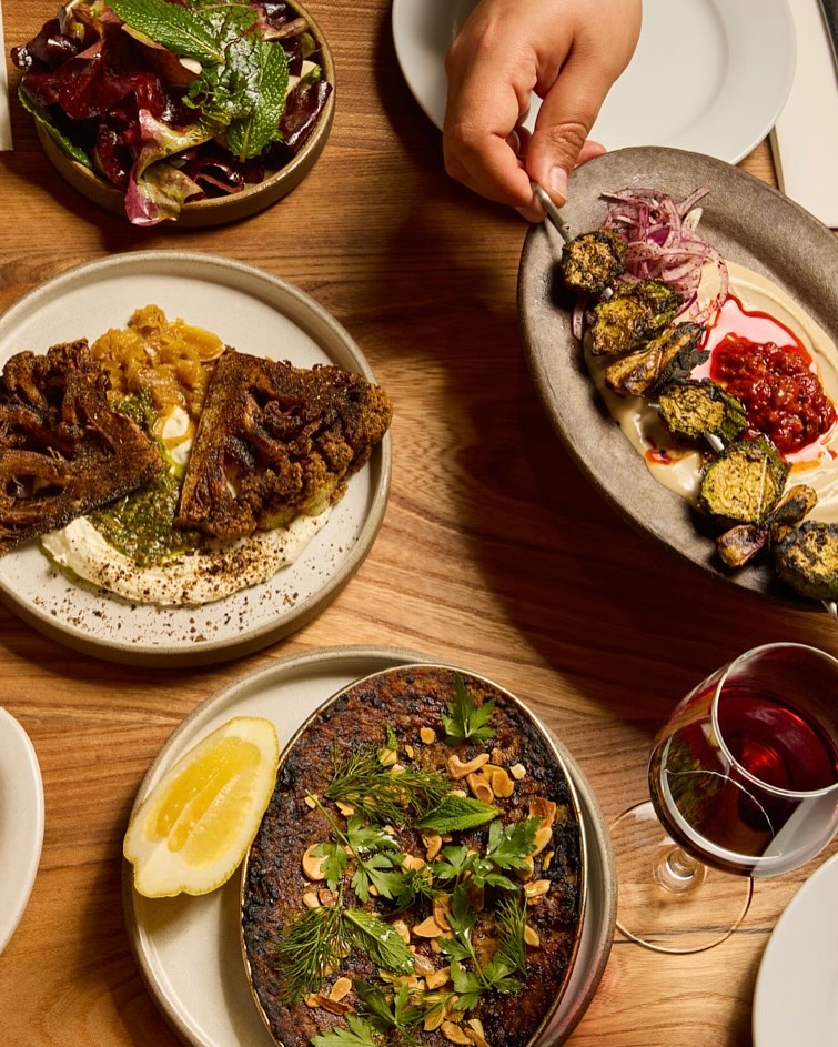 A person serving a plate of food including grilled brussels sprouts, hummus, and a red sauce.