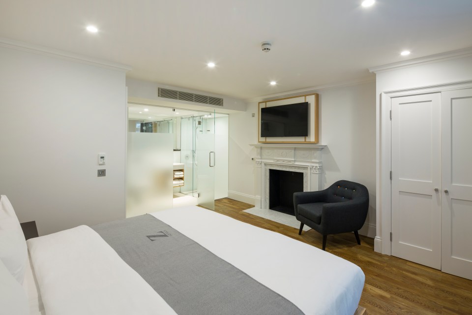 A contemporary bedroom at Z Gloucester Place, with a king-sized bed, fireplace, armchair, and a glass-enclosed bathroom.