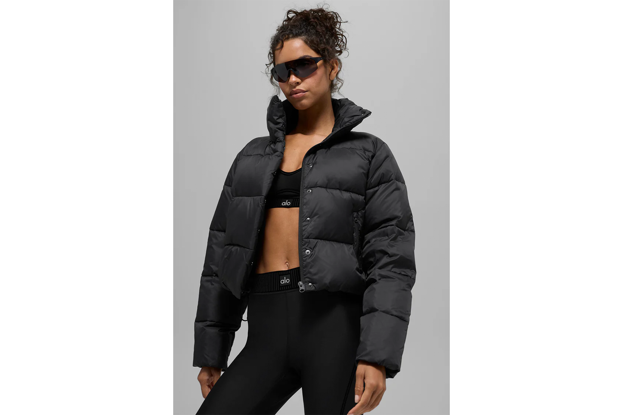 Woman wearing a black puffer jacket, Alo sports bra, Alo leggings, and sunglasses.