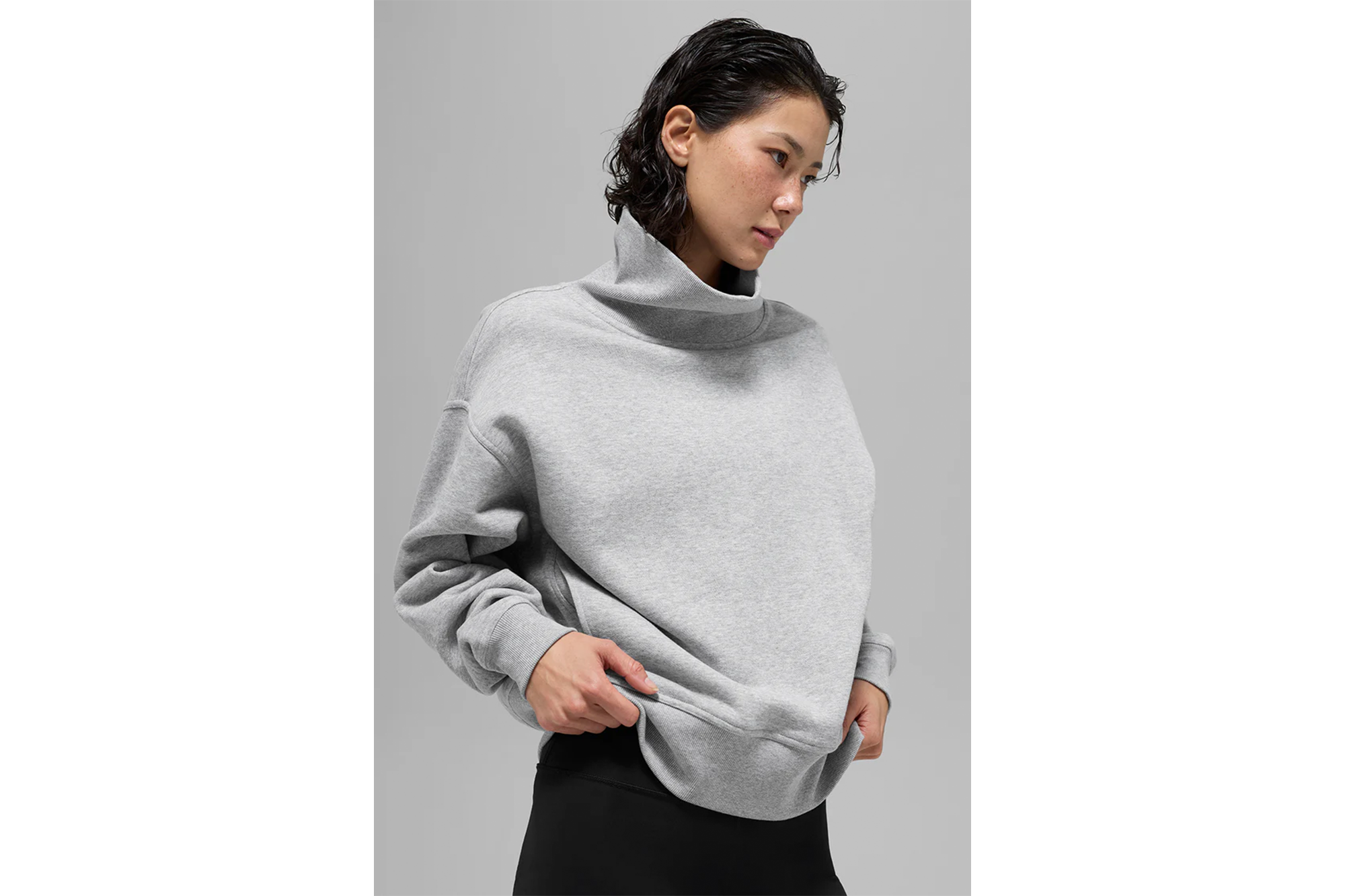A woman wearing a gray funnel neck sweatshirt and black leggings, pulling the hem of the sweatshirt.