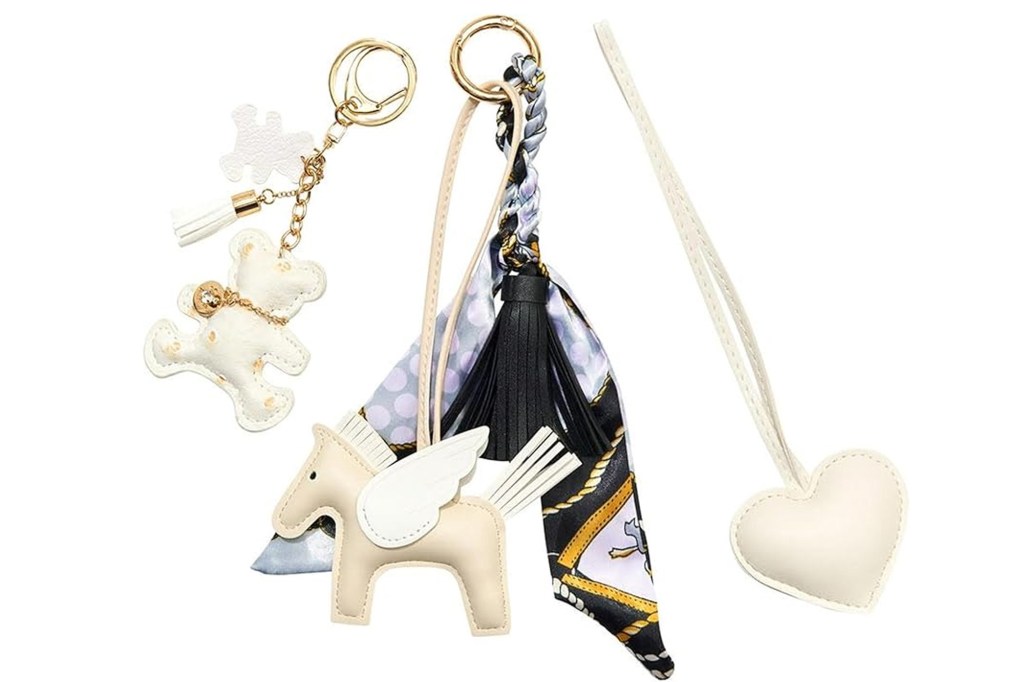 Three bag charms including a white teddy bear, a white Pegasus with a patterned scarf and black tassel, and a white heart.