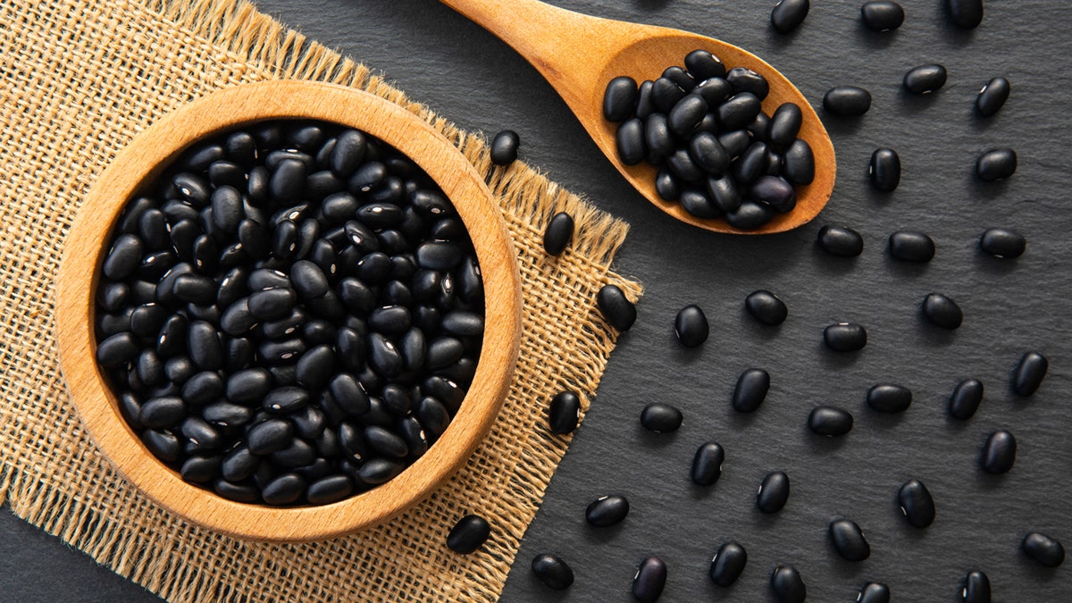 Dried raw black beans in a wooden bowl, spoon and scattered on a table.