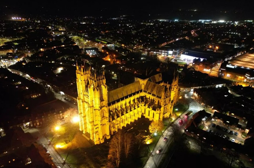 Aerial night view of a large, illuminated cathedral surrounded by a city with scattered lights.