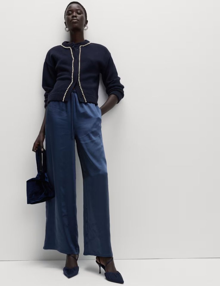 A model wearing a navy blue cardigan with pearl detailing, blue satin wide-leg trousers, and holding a navy velvet handbag.