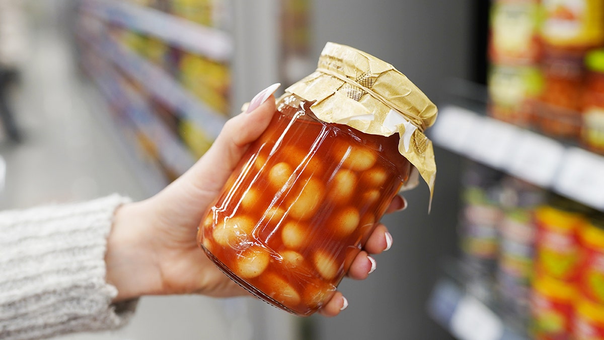 Woman's hand holding glass jar of beans in tomato sauce in grocery store.