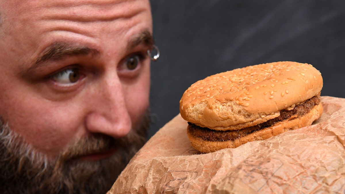 Casey Dean inspecting a McDonald's burger bought in 1995, seen staring at burger up-close, with beard and eyebrow ring.