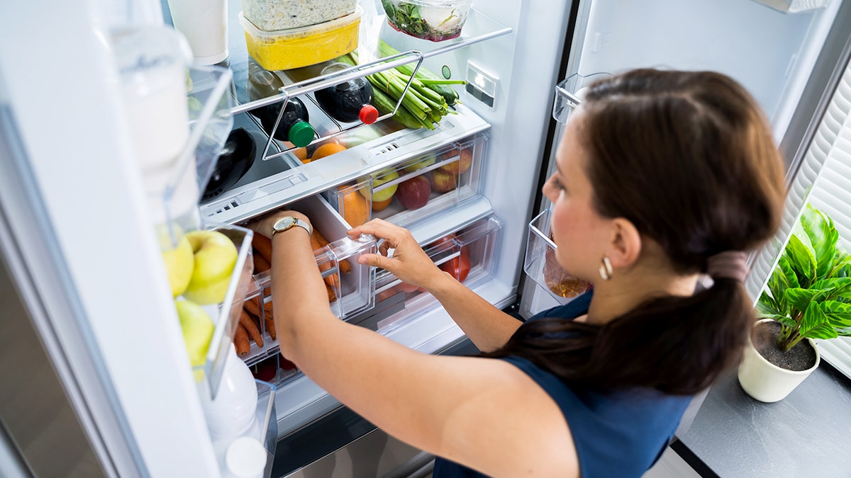 A woman wearing a watch reaches for carrots inside a produce drawer in a refrigerator.